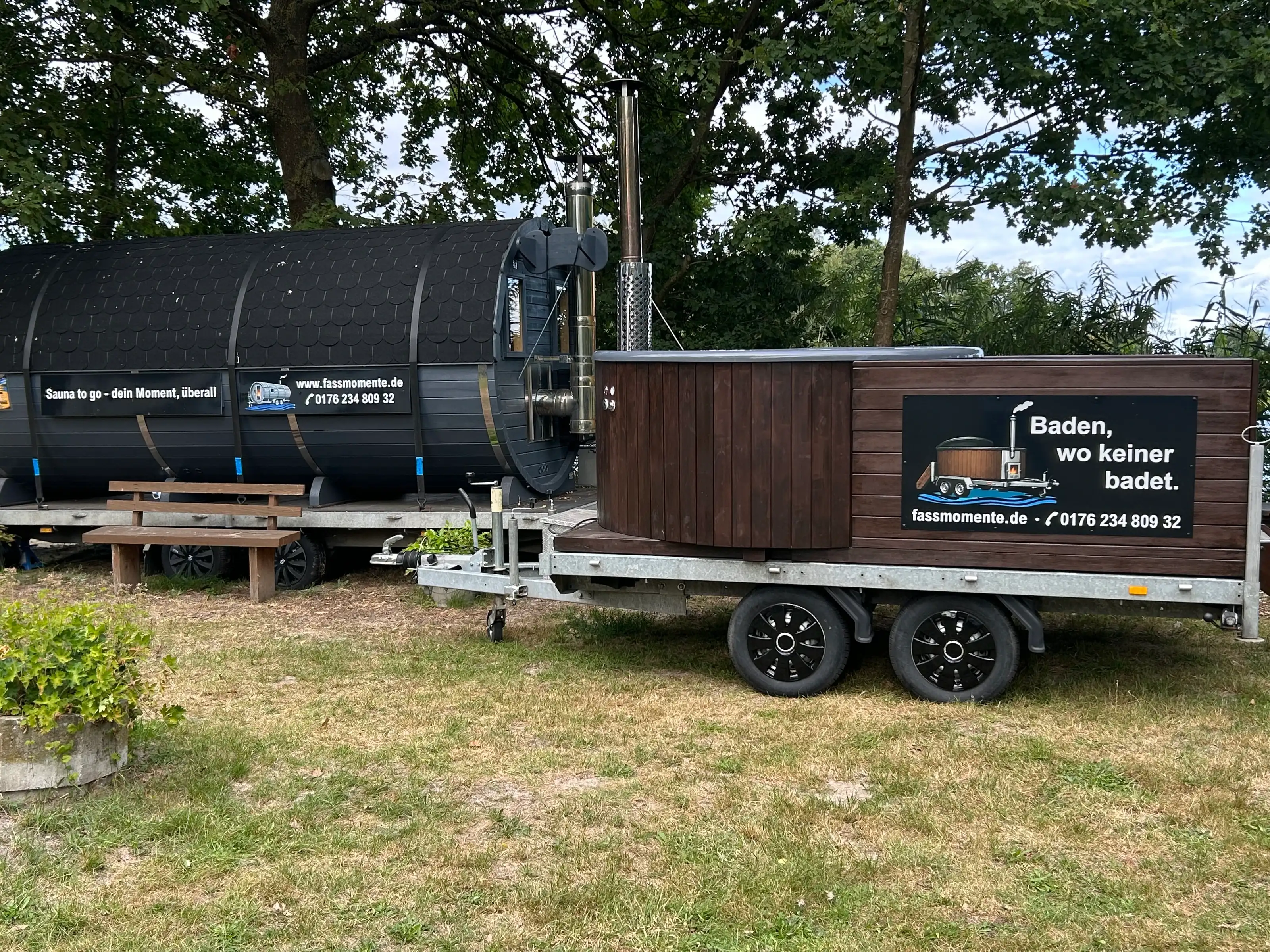 Fasssauna und Whirlpool auf ihren Anhängern nebeneinander auf einer Wiese