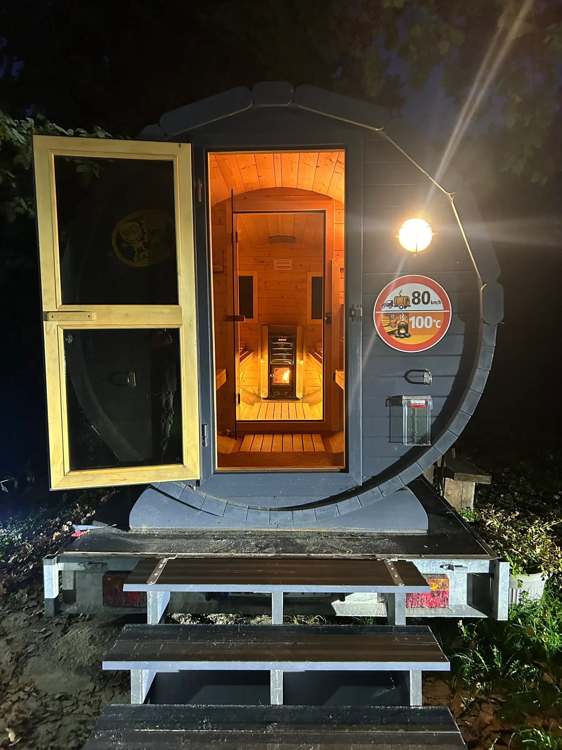 Fasssauna am Abend mit offener Außentür, Treppe und Außenleuchte, hinten brennt der Holzofen
