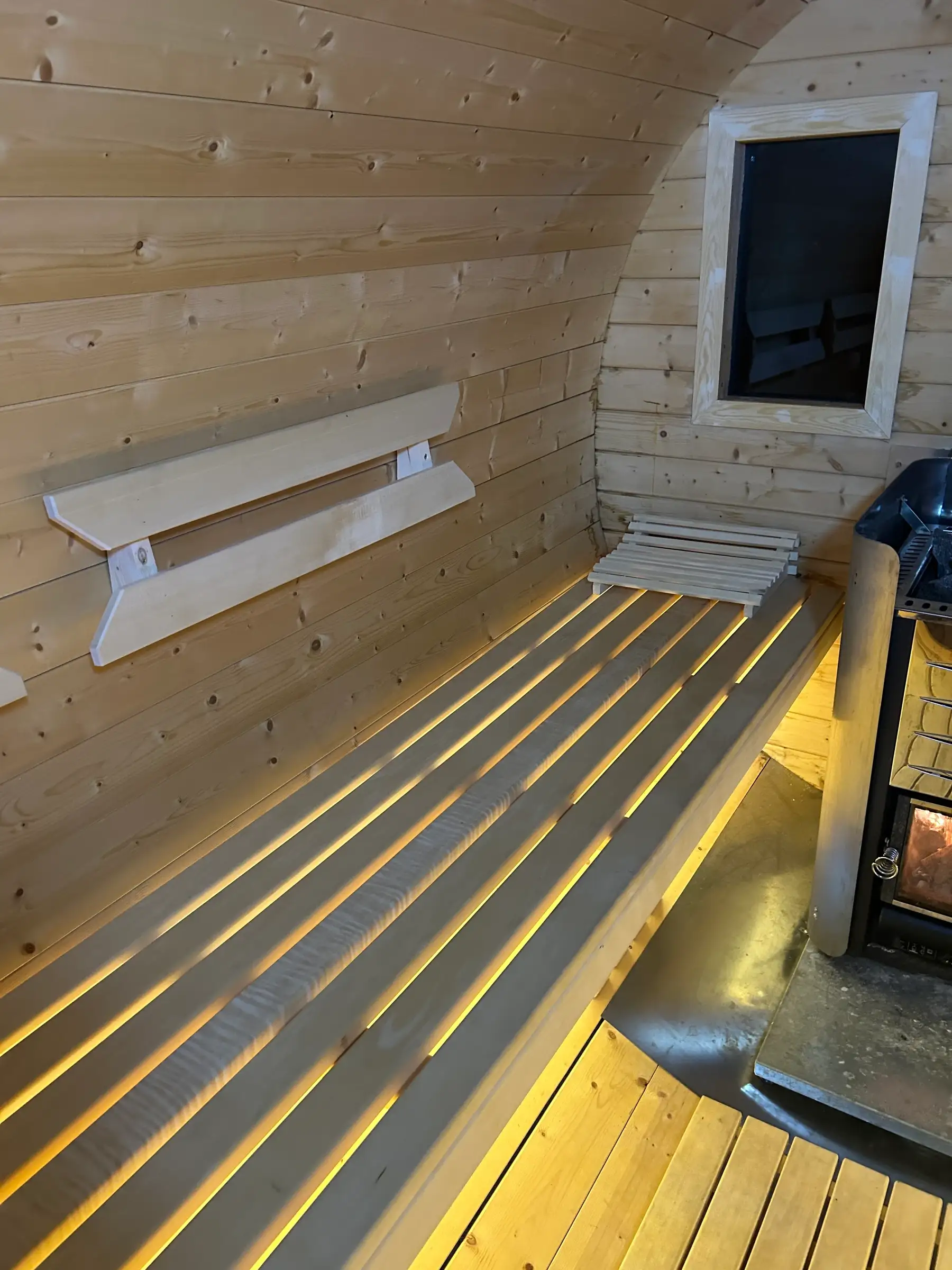 Liegebank aus hellem Holz mit Kopfstütze und Rückenlehne, rechts der Holzofen