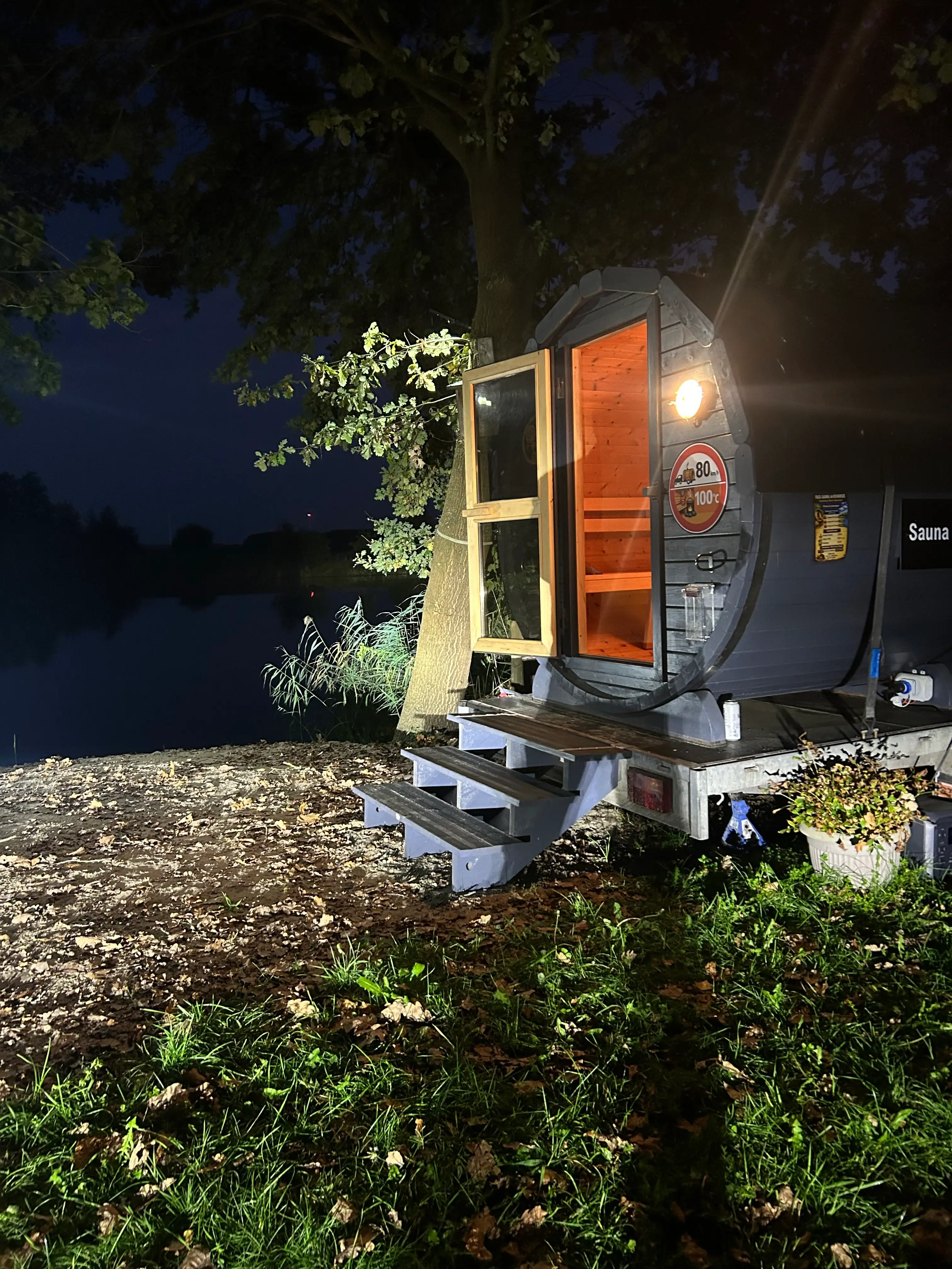 Fasssauna am Reihersee bei Lüneburg am Abend, die Tür steht offen und der Saunaraum leuchtet