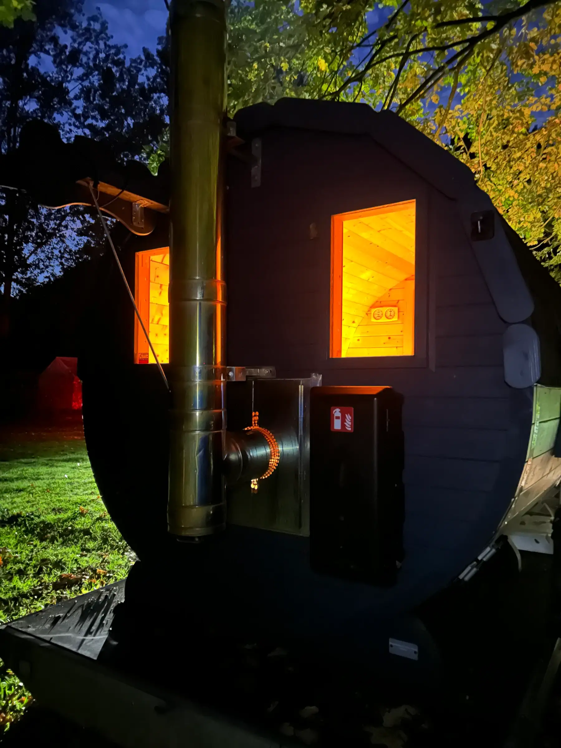 Rückseite der Fasssauna bei Nacht mit Edelstahlschornstein, warm leuchtenden Fenstern und Kasten für den Feuerlöscher